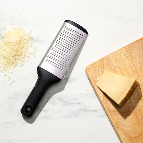 An OXO stainless steel handheld grater grating cheese onto a cutting board.
