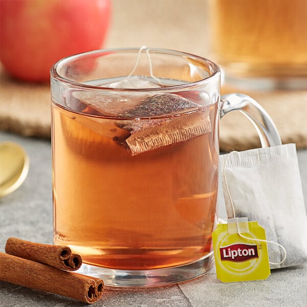 A glass mug of Lipton Cinnamon Apple Herbal Tea with a tea bag and cinnamon sticks.