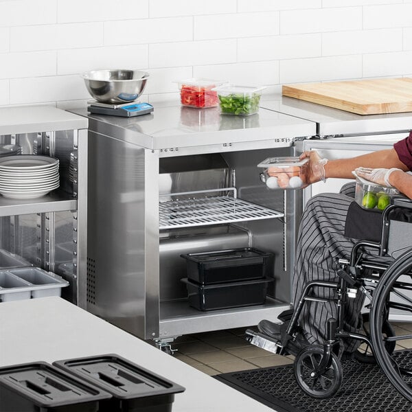 A person in a wheelchair holding a container of food with green vegetables in a stainless steel undercounter refrigerator.