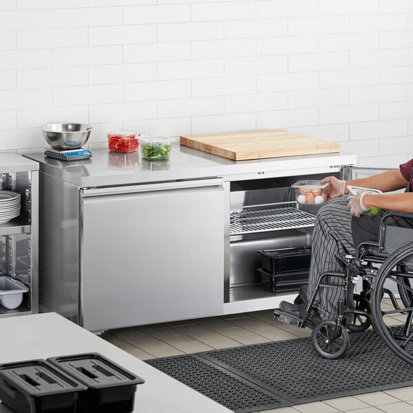 A woman in a wheelchair using an Avantco stainless steel undercounter refrigerator.