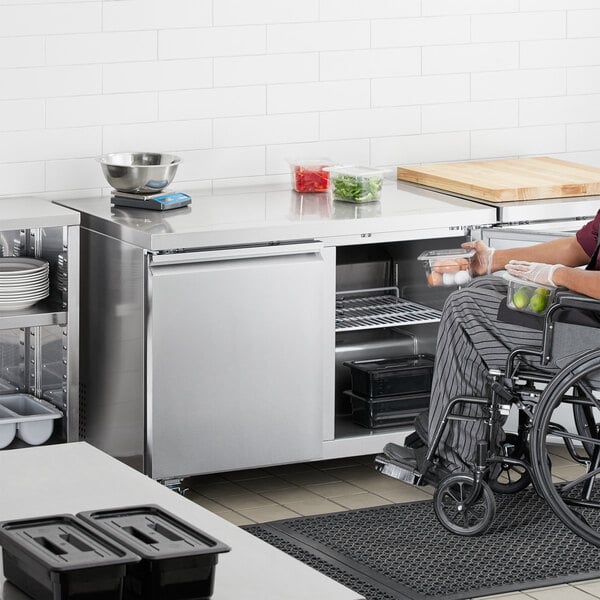 A woman in a wheelchair using an Avantco stainless steel undercounter refrigerator in a kitchen.