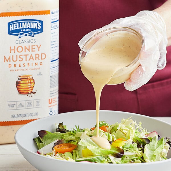 A person pouring Hellmann's honey mustard dressing into a bowl of salad.