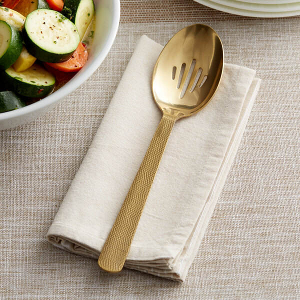 A gold slotted serving spoon on a white napkin next to a bowl of vegetables.
