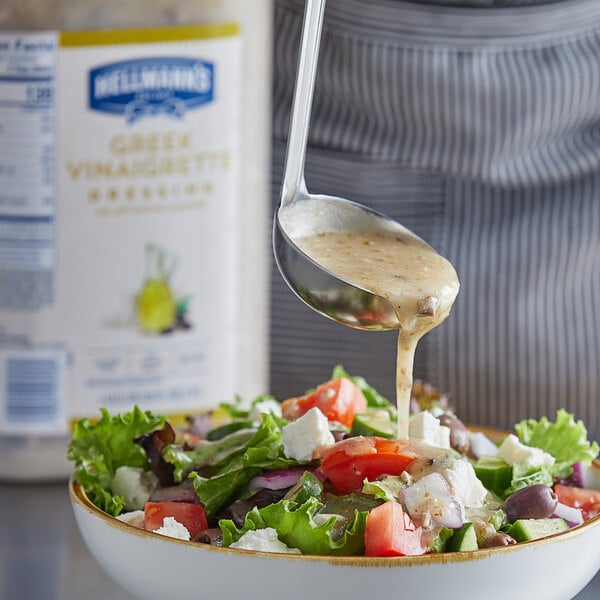 A spoon pouring Hellmann's Greek Vinaigrette dressing into a bowl of salad.