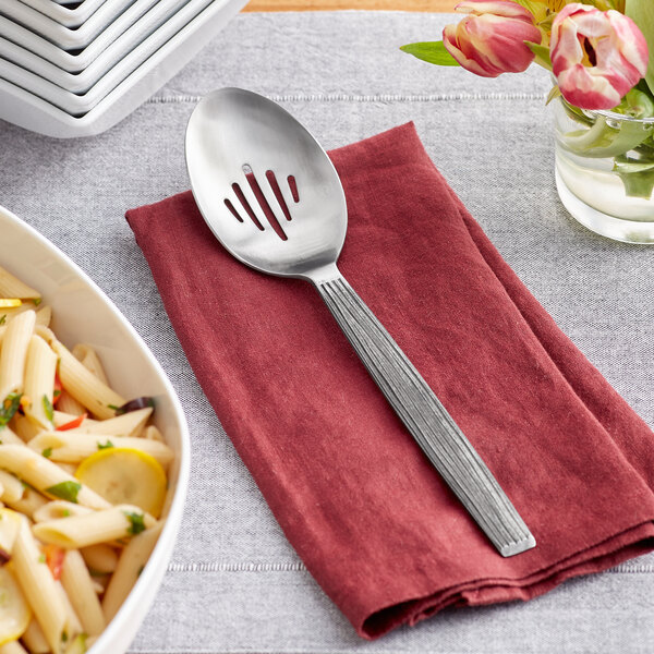 An American Metalcraft stainless steel slotted spoon on a napkin next to a bowl of pasta and a glass of water