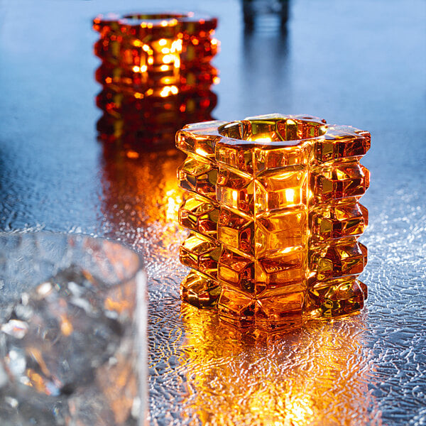 A Hollowick amber glass faceted cube votive with a lit candle inside.