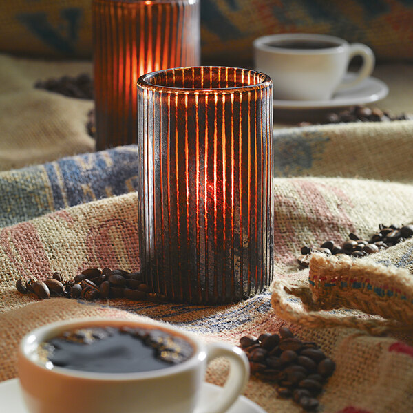A Hollowick mocha glass cylinder candle holder with a lit candle and coffee beans on a table.