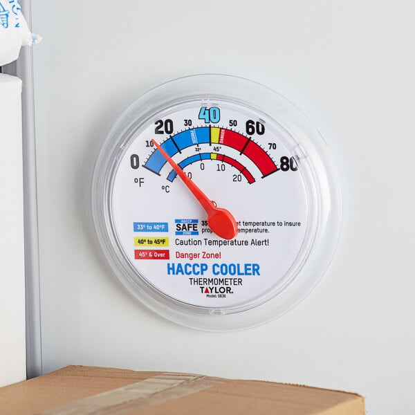 A Taylor HACCP wall thermometer with a red needle on a white wall.