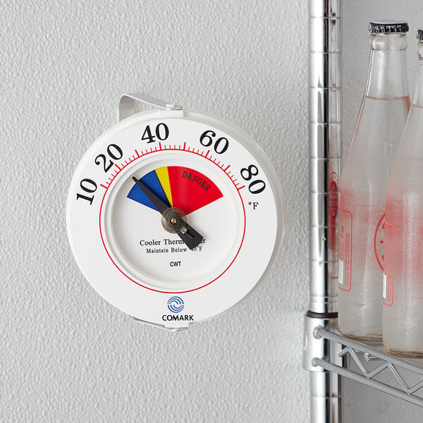 A Comark HACCP cooler wall thermometer with a white dial on a wall next to bottles of liquid.