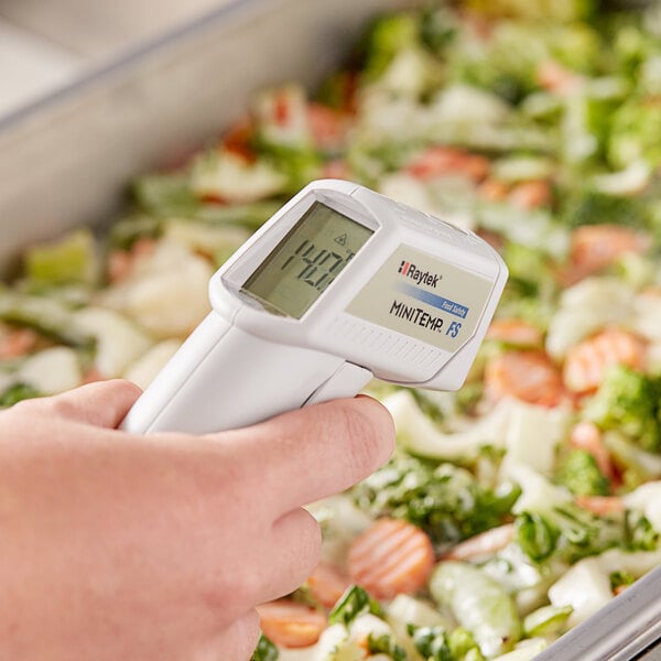 A hand using a Comark infrared thermometer to measure the temperature of a bowl of vegetables.