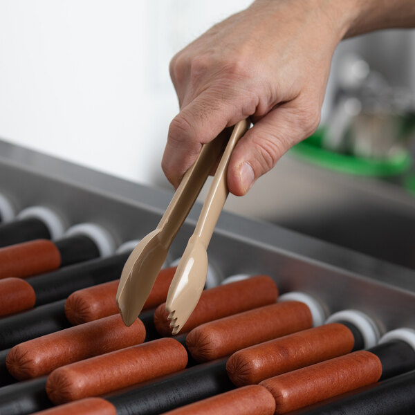A person holding APW Wyott plastic tongs over hot dogs.