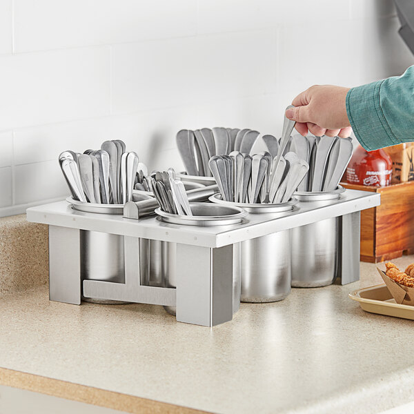 A hand holding a spoon in a stainless steel tray with 5 other cylinders filled with silverware.