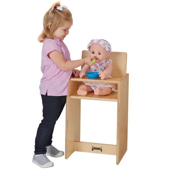 A girl standing next to a table feeds a baby doll in a wood high chair.