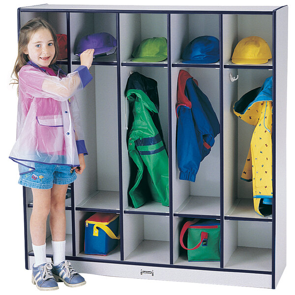 A young girl in a plastic raincoat standing next to a Rainbow Accents navy coat locker with green and red bags.