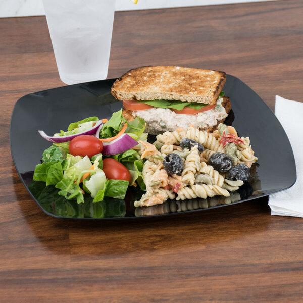 A Fineline black plastic plate with a sandwich and salad on a table.