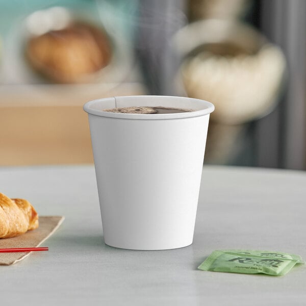 A white Choice paper hot cup filled with brown liquid on a table with a croissant.