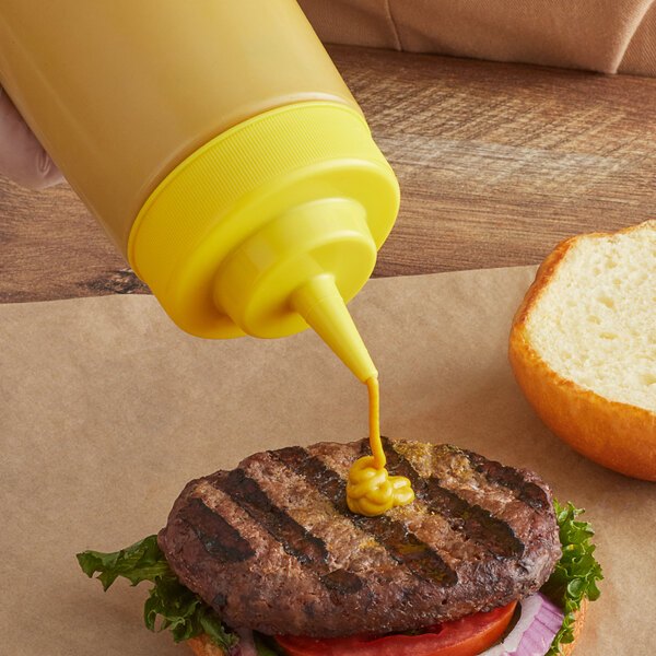 A hand using a Tablecraft solid yellow widemouth cone tip cap to pour mustard onto a hamburger.