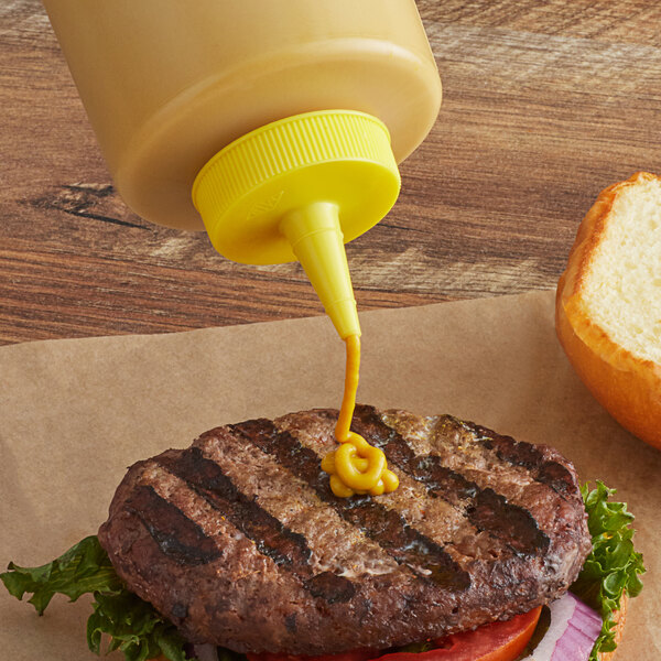A Tablecraft yellow plastic squeeze bottle cap being used to pour mustard onto a burger.