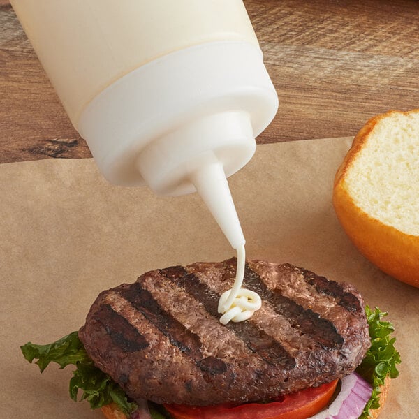 A sauce being poured from a white Tablecraft squeeze bottle with a clear cone tip cap onto a burger.