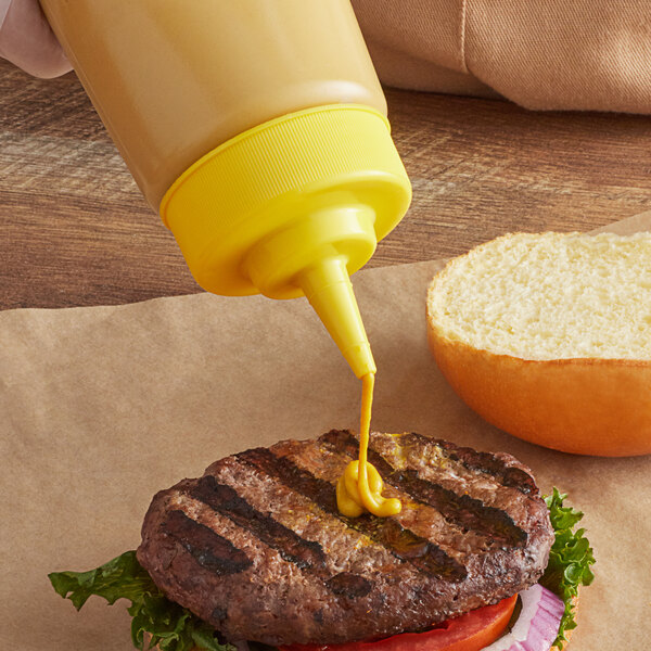 A hand pours yellow mustard from a Tablecraft Widemouth Cone Tip Cap onto a burger.