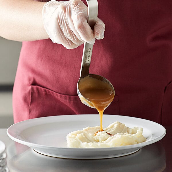 A person using a Vollrath stainless steel ladle to pour liquid on a plate of mashed potatoes.