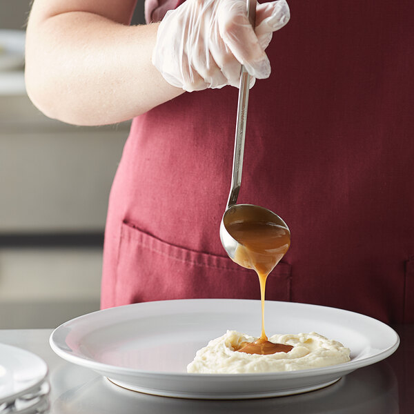 A person using a Vollrath stainless steel ladle to pour brown liquid on a white plate.