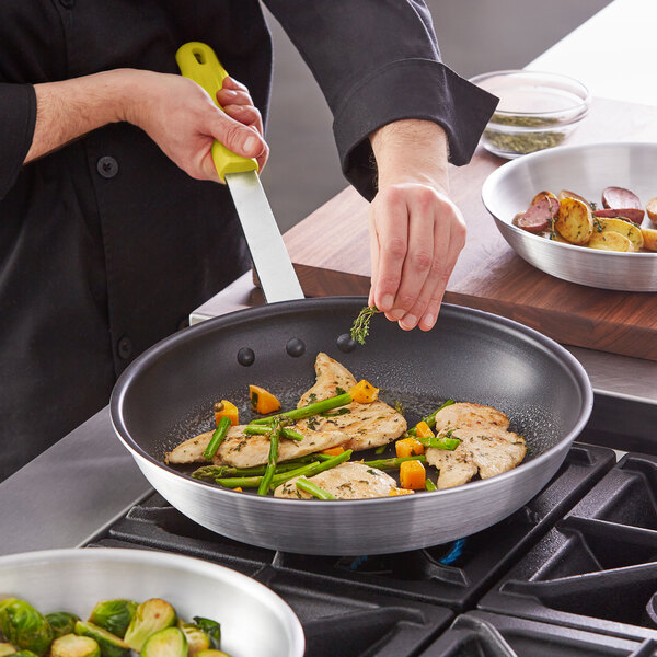 A person cooking food in a Choice aluminum non-stick fry pan with a yellow silicone handle.