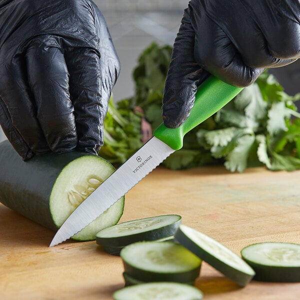 A person in black gloves using a Victorinox green paring knife to cut a cucumber.