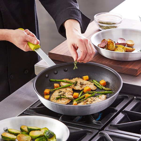 A person cooking food in a Choice aluminum non-stick fry pan with a yellow silicone handle.