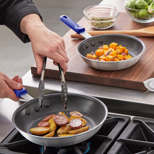 A person cooking potatoes and sausages in a Choice aluminum non-stick frying pan with blue silicone handles.