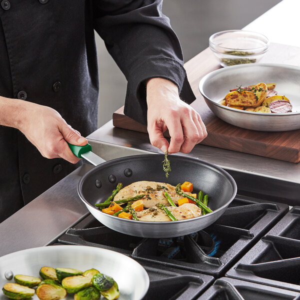 A person cooking food in a Choice aluminum non-stick fry pan with a green silicone handle.