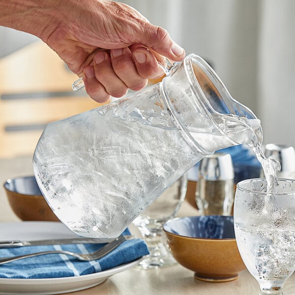 A hand pouring water from a glass into an Acopa glass pitcher.