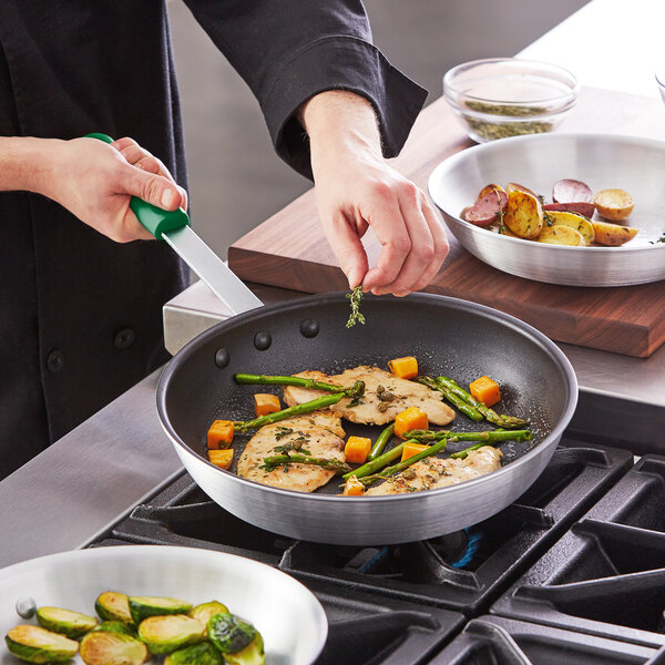 A person cooking food in a Choice aluminum non-stick fry pan with a green handle.