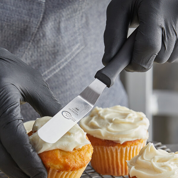 A person in black gloves using a Mercer Culinary offset icing spatula to spread frosting on cupcakes.