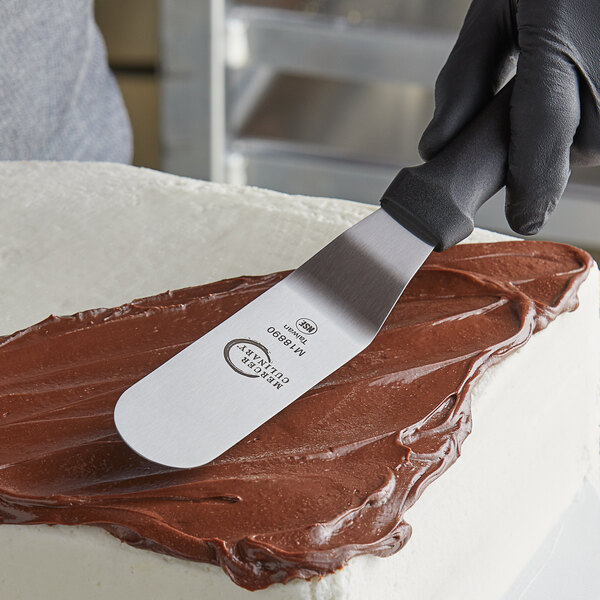 A person using a Mercer Culinary offset spatula to frost a cake with chocolate frosting.