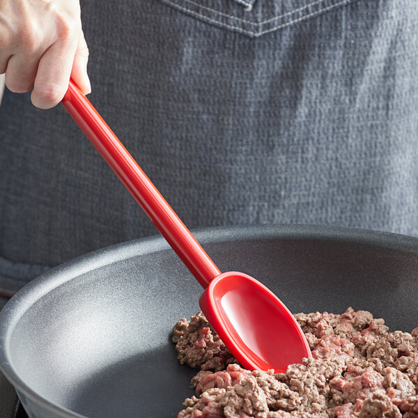 A hand using a red Mercer Culinary Hell's Tools spoon to stir food in a pan.