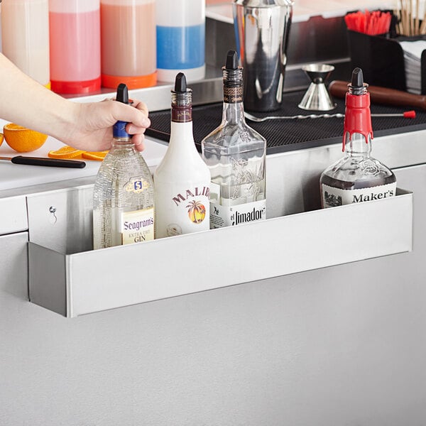 A person uses a Regency stainless steel single tier speed rail to hold bottles of liquor on a counter.