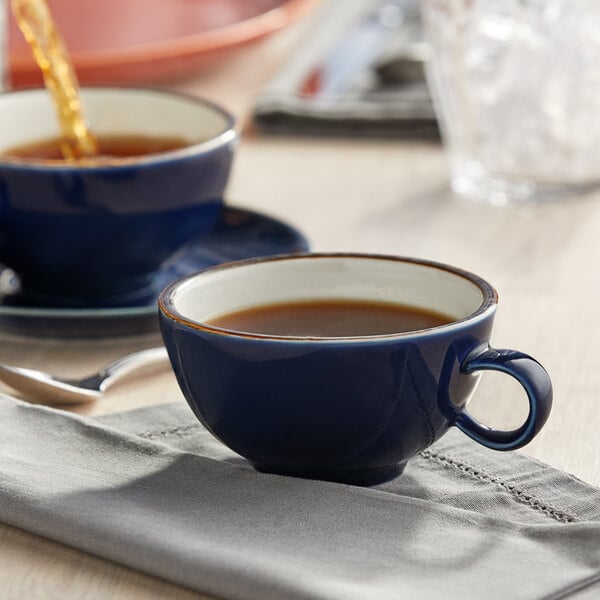 A blue Acopa Keystone stoneware cup filled with brown liquid on a saucer.