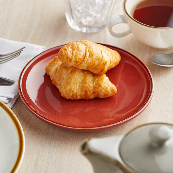 A Acopa Keystone Sedona Orange stoneware coupe plate with a croissant and tea on a table.