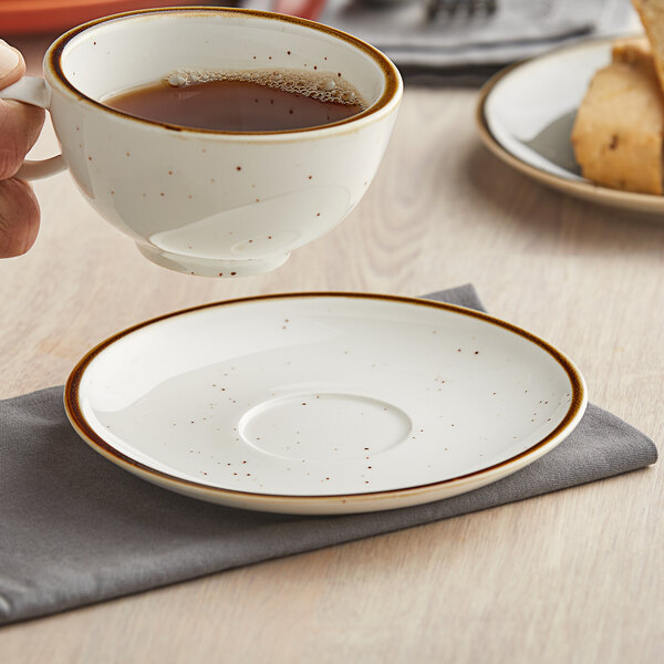 A hand holding a cup of coffee over a white Acopa Keystone Vanilla Bean saucer with brown speckles.