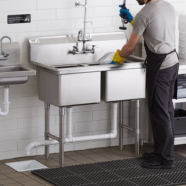 A man in an apron and gloves using a Regency 2 compartment stainless steel sink in a professional kitchen.