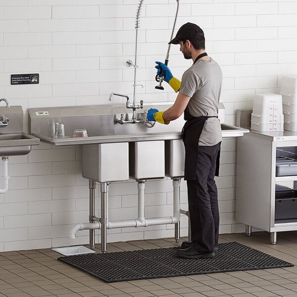 A man in a uniform and gloves washing a Regency stainless steel 3 compartment sink.