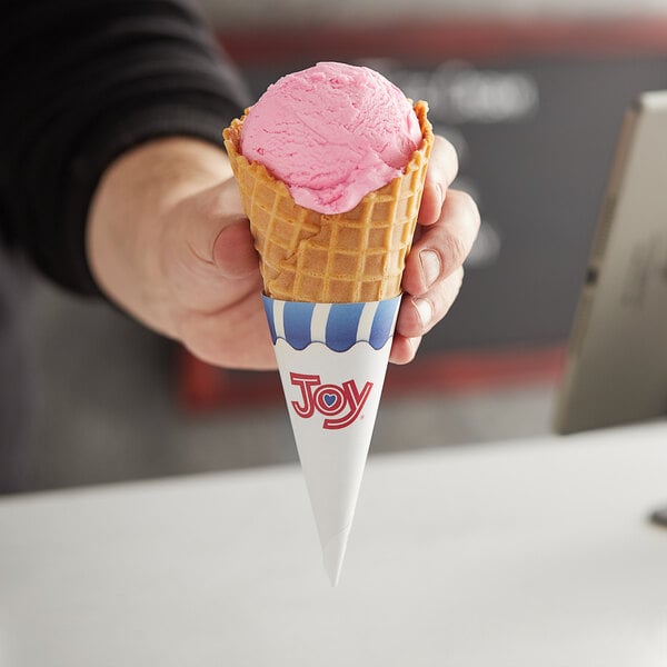 A hand holding a JOY large waffle cone with a scoop of pink ice cream.
