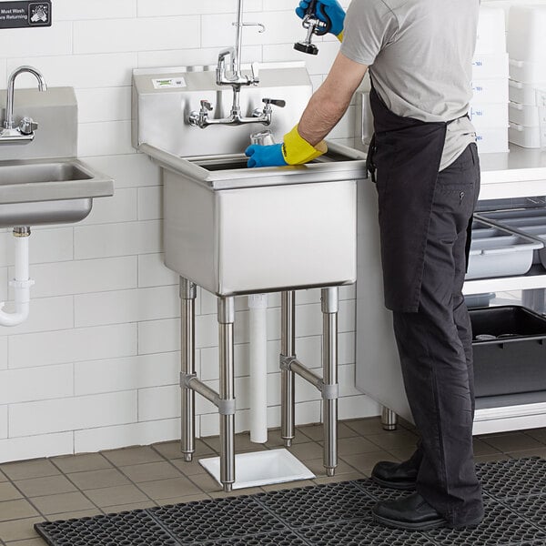 A man in yellow gloves standing in a Regency stainless steel one compartment sink in a professional kitchen.