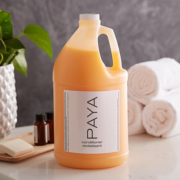 A jug of PAYA Papaya conditioner on a counter next to towels and a plant.