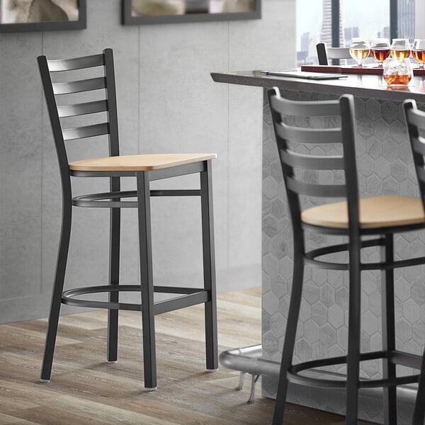 A Lancaster Table & Seating black ladder back bar stool with a natural wood seat next to a table.