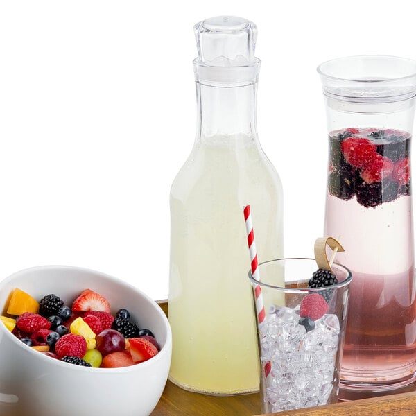 A Tablecraft Swirl plastic carafe with a glass of liquid and a bowl of fruit on a table.
