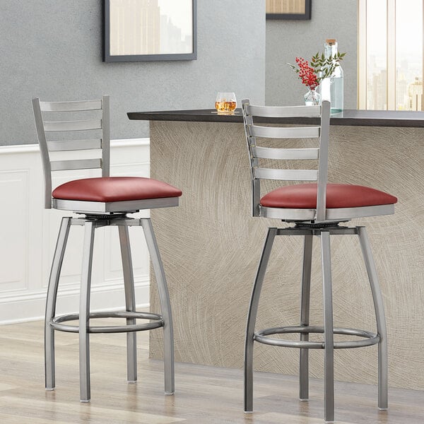 Two Lancaster Table & Seating clear coat finish ladder back swivel bar stools with burgundy vinyl padded seats at a silver countertop.