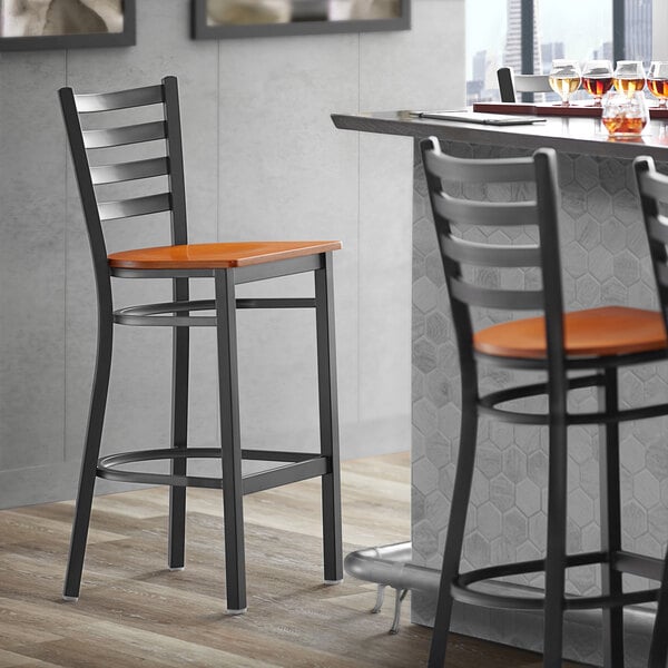 A Lancaster Table & Seating black finish ladder back bar stool with a cherry wood seat next to a table.