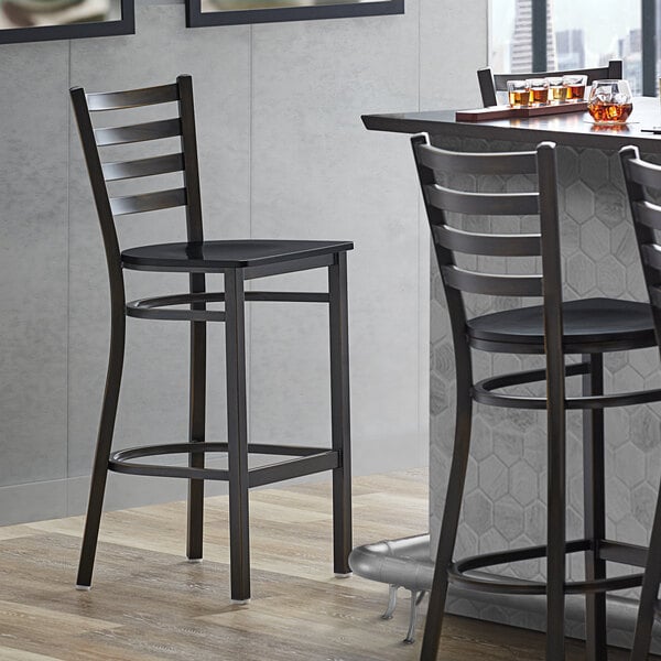 A Lancaster Table & Seating black wood ladder back bar stool with a copper finish next to a table.